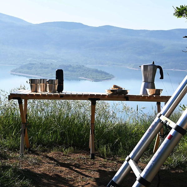 Table en bois pliable pour le camping avec vaisselle sur fond de lac et de montagnes, idéale pour les repas en plein air, robuste et compacte.
